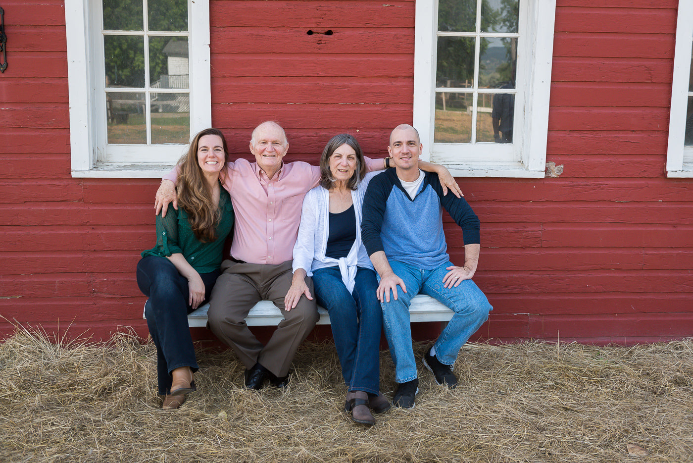 The family behind Bon Bee Honey sitting on a hay bale in front of a red building with white windows.