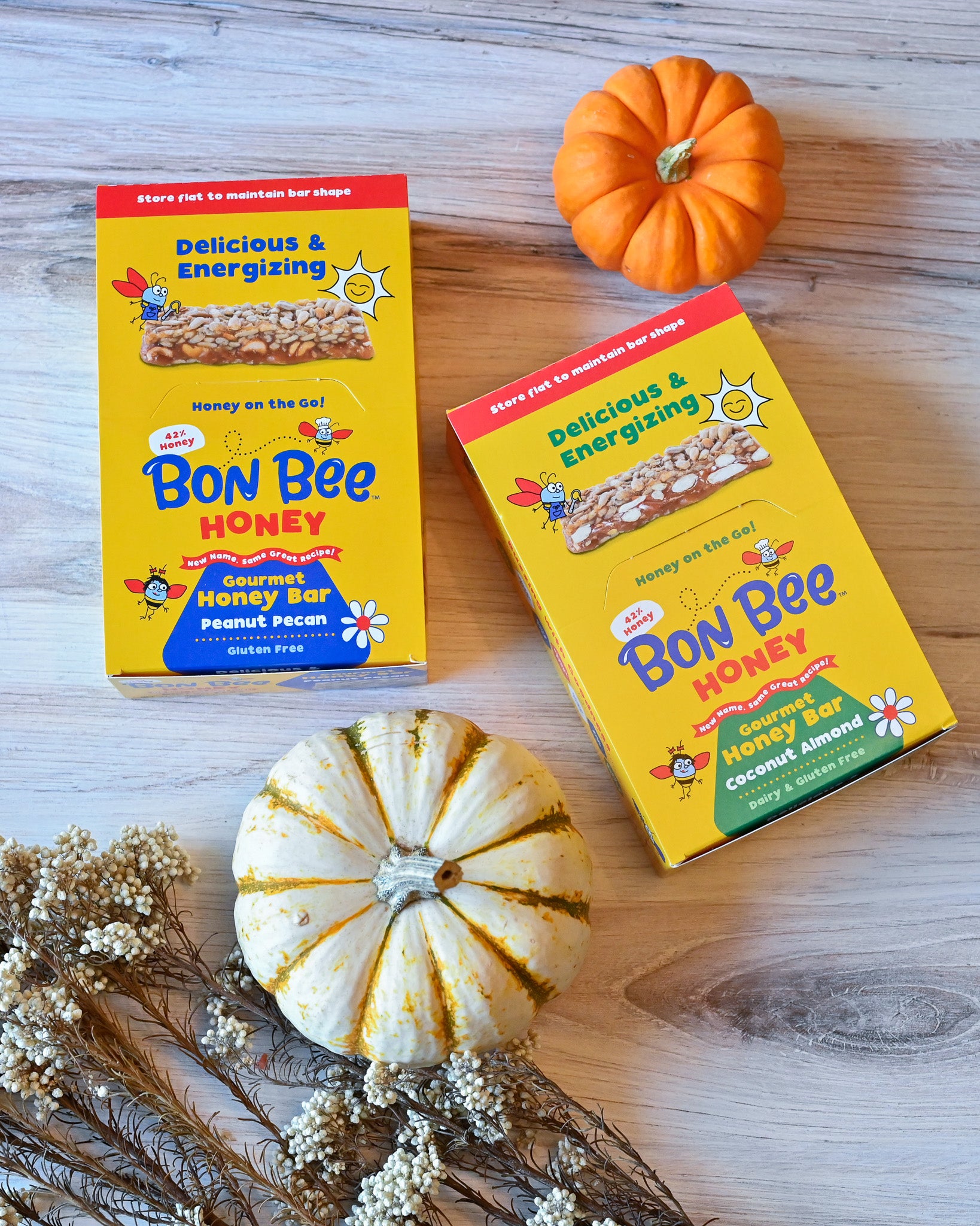Two boxes of Bon Bee Honey bars on a wooden surface with small pumpkins and flowers.